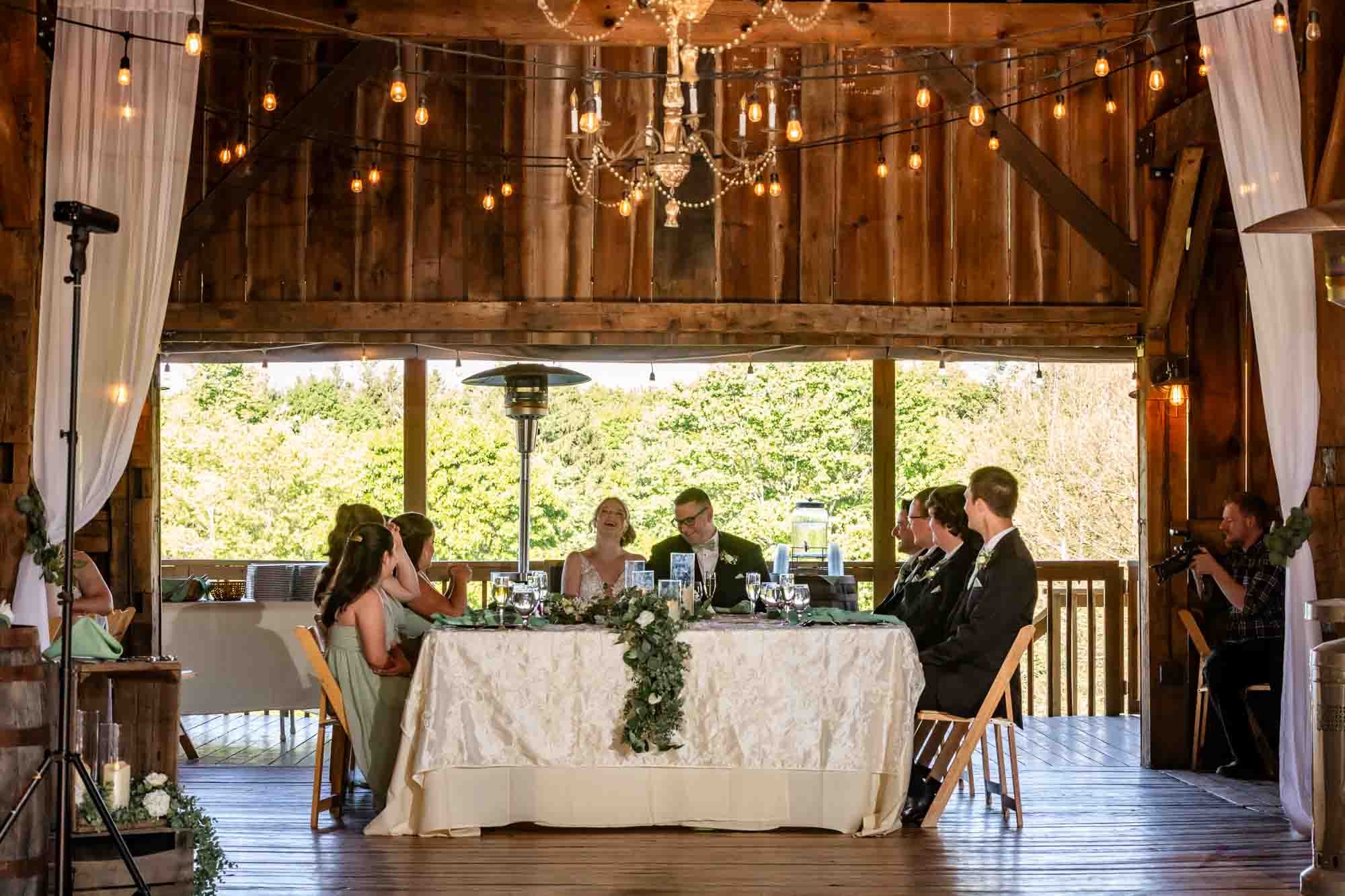 bride and groom sit at a sweetheart table surrounded by their bridal party in their barn reception at pinehall at eisler farms